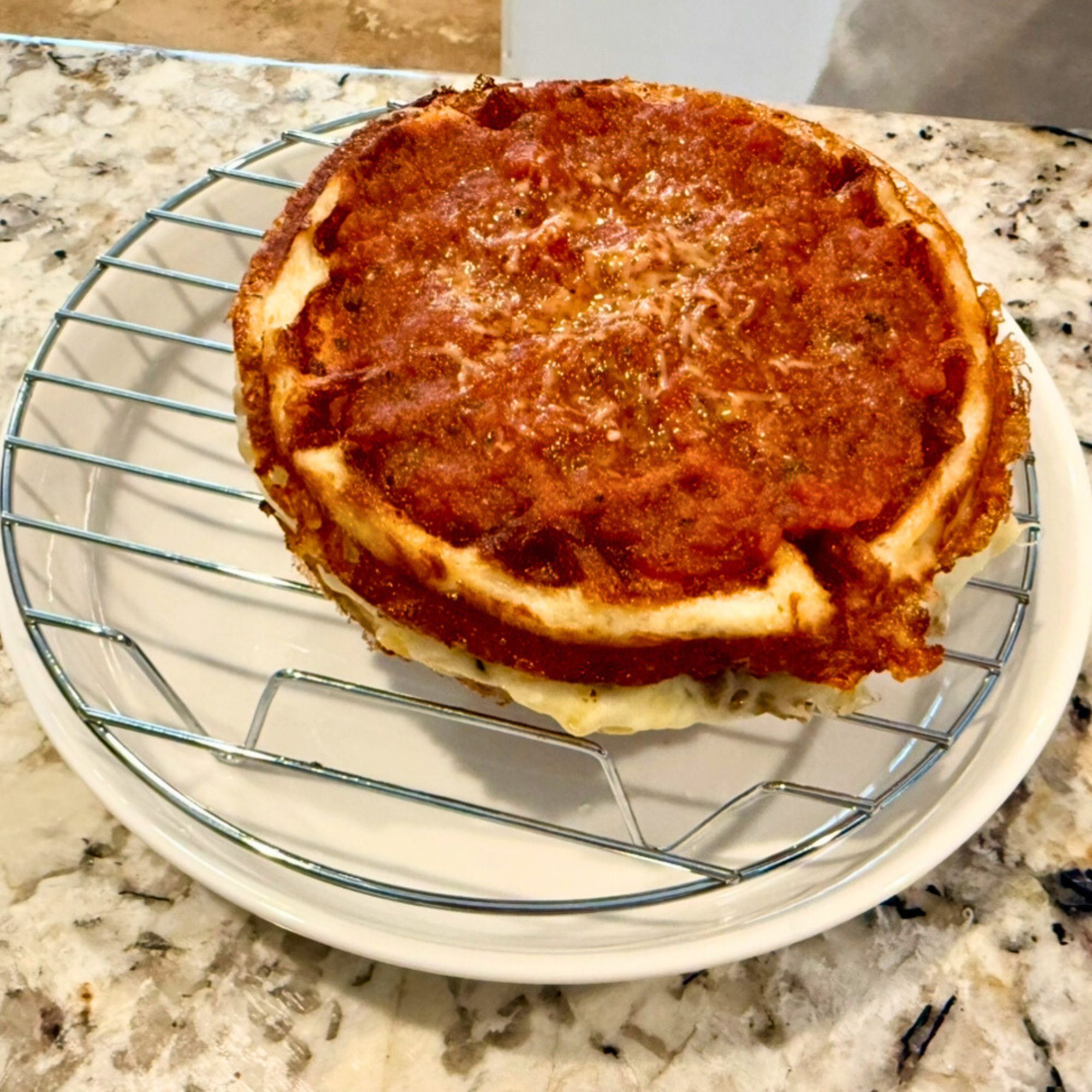 Golden brown keto pizza with marinara and melted cheese, cooling on a wire rack over a white plate on a granite countertop.