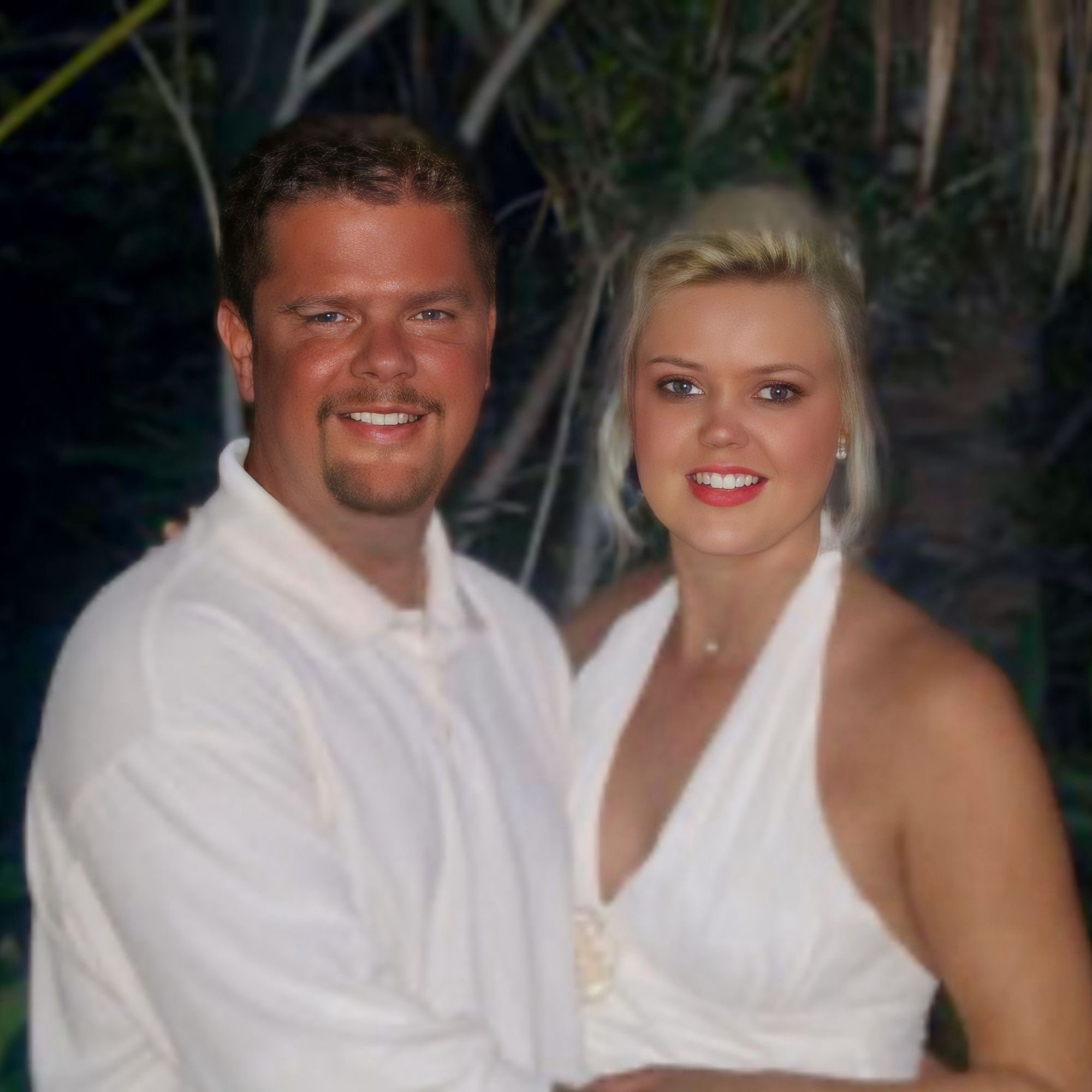 Smiling bride and groom in white attire embracing under moonlight on their wedding night, surrounded by soft tropical greenery.