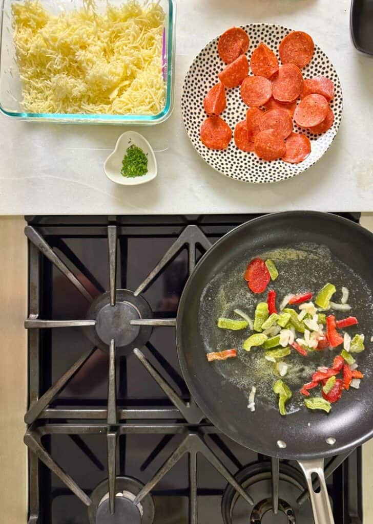 Overhead view of shredded cheese, sliced pepperoni, and a skillet of sautéing peppers and onions — ingredients prepped for making a keto pizza omelet.