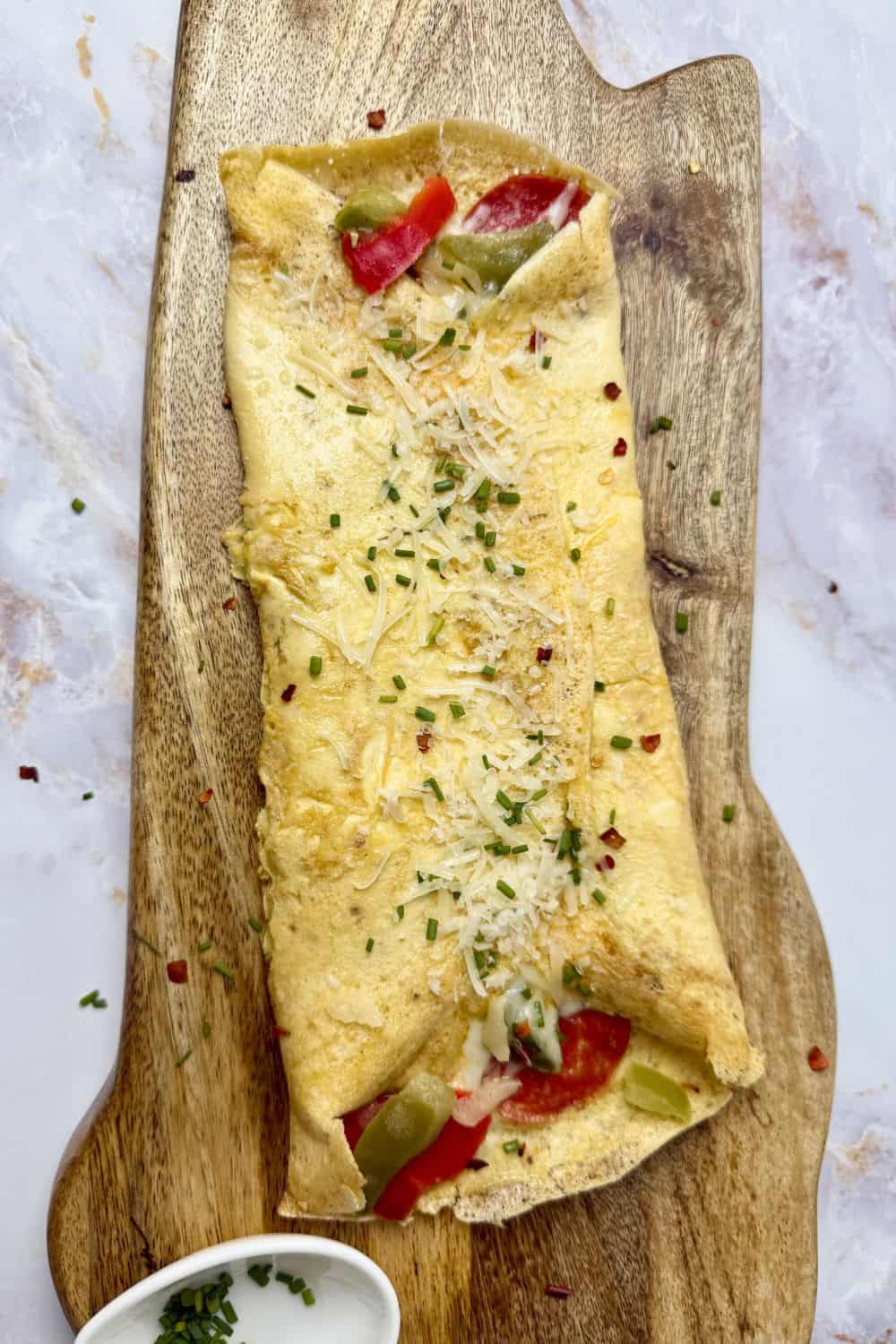 Golden omelet folded over melted cheese, savory pepperoni, and sautéed red and green peppers with onion, garnished with herbs and served on a wooden board with dipping sauce.