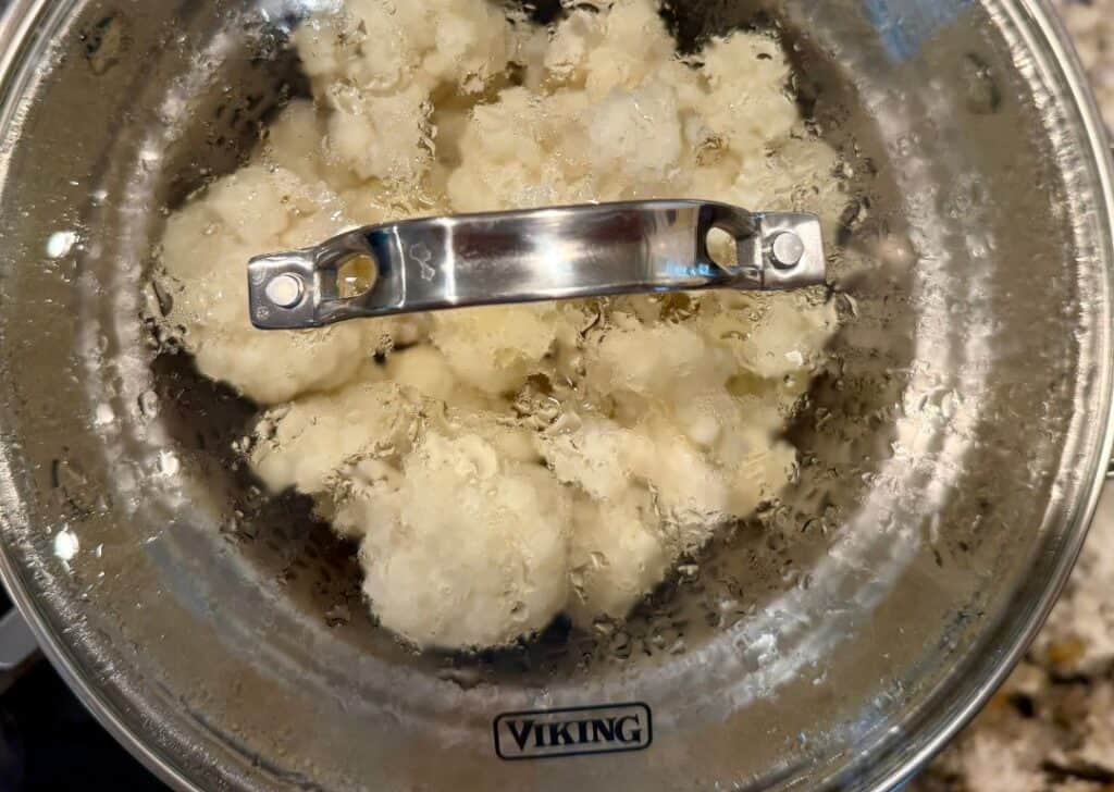 Cauliflower florets steaming in a covered stainless steel pot to make creamy keto cheesy grits.