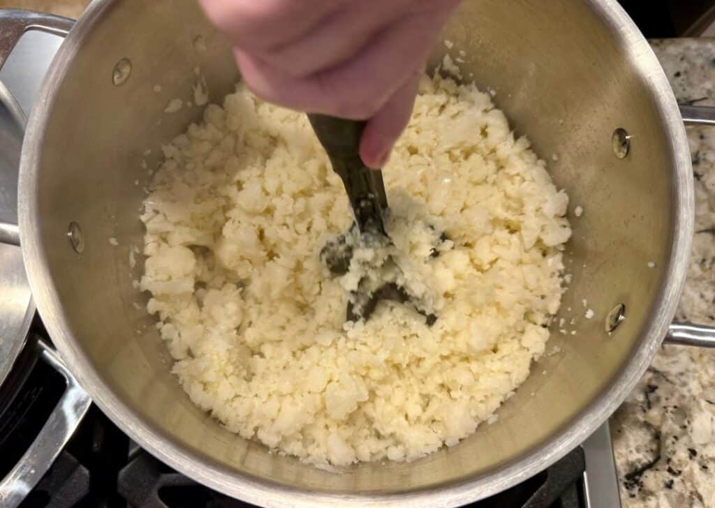 Mashing steamed cauliflower in a pot to make chunky, creamy keto cheesy grits.