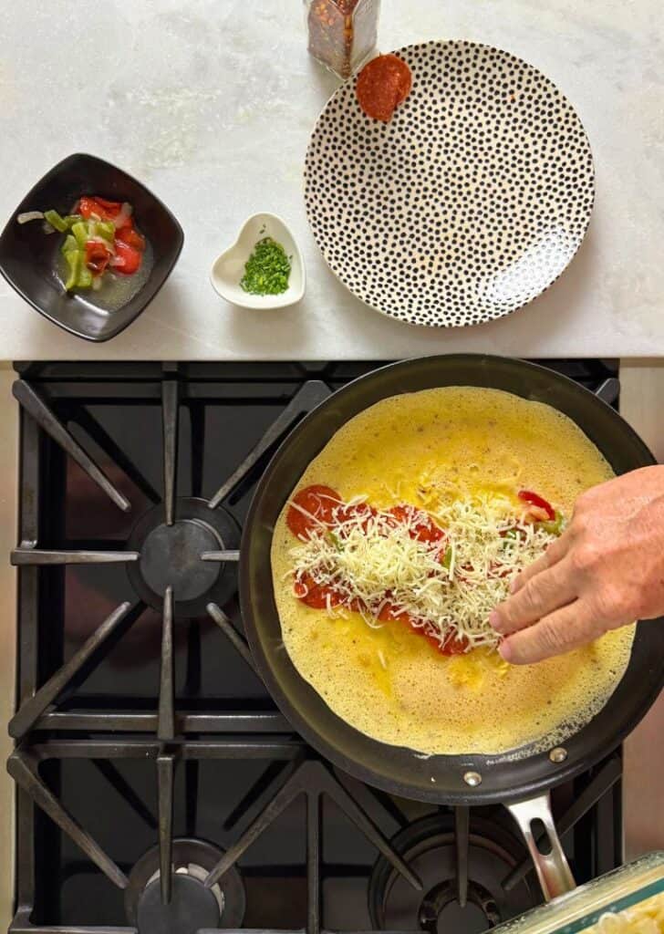 Hand adding shredded cheese and pepperoni onto an omelet cooking in a skillet, with toppings staged above on the counter.